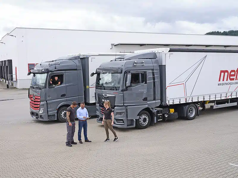 Zwei graue LKW vor Lagerhalle mit Personen